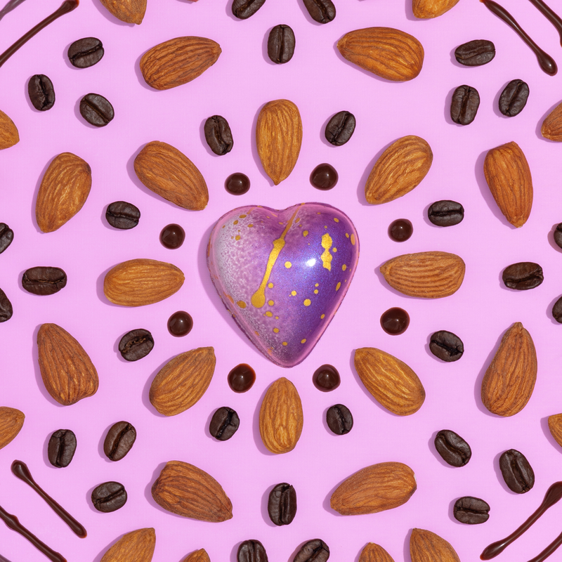 A beautiful purple Mocha Almond heart-shaped truffle sitting on a purple background. The truffle is placed in the center with almonds, drops of chocolate syrup and espresso beans placed in a circular pattern to accentuate the chocolate.  
